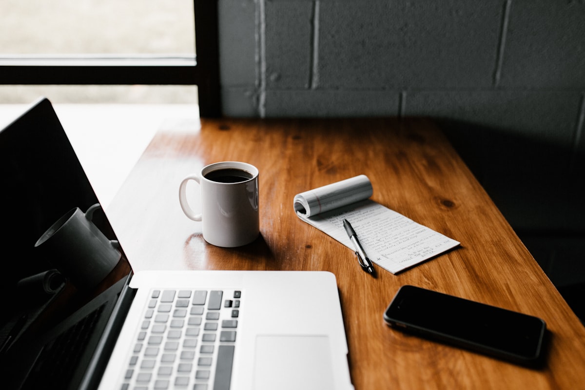 A laptop, coffee mug, notebook, and phone arranged on a desk, representing a corporate gift phone case setup